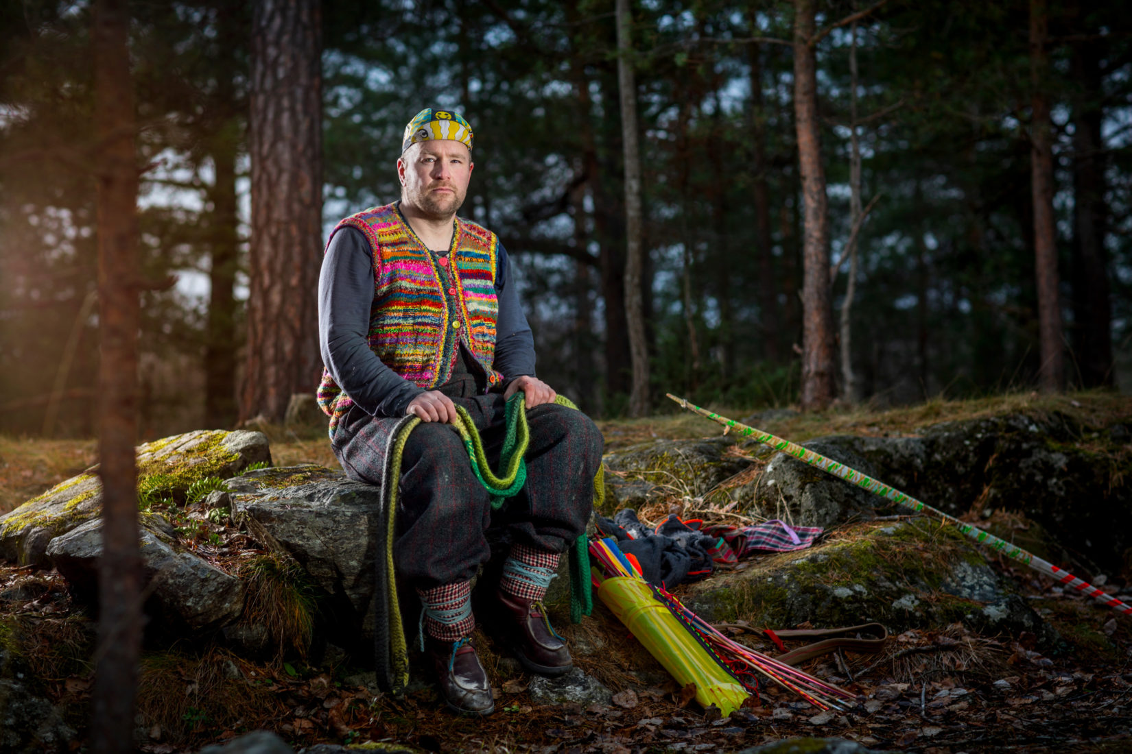 Anders Jakobsen, designer, assis sur un rocher dans la forêt suédoise tenant un arc en bois dans sa main.