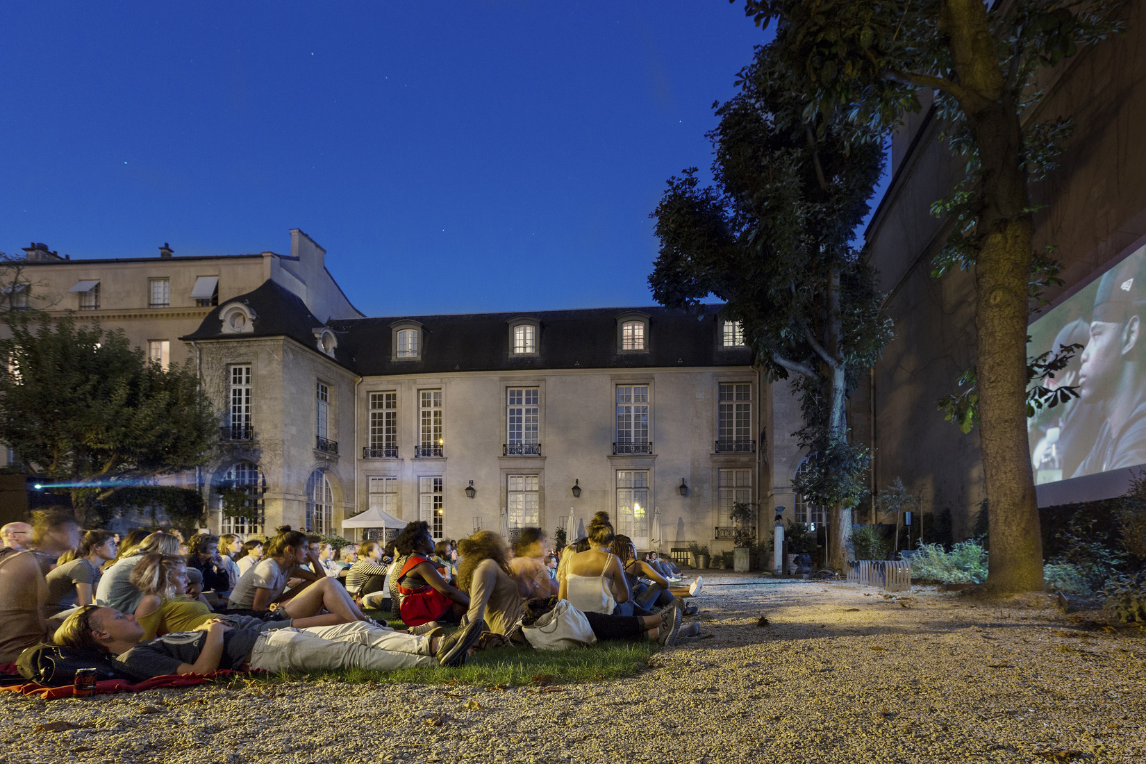 A big group of people sit in the garden of Hôtel de Marle during an outdoor cinema session.