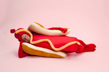 Un homme allongé, déguisé en hotdog, fond rose