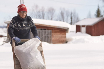 Maj Doris de Jom Jon Blåhed. Le format de l'image a été modifié. une vieille femme sami pousse un traineau dans la neige - on voit quelques maisons en fond