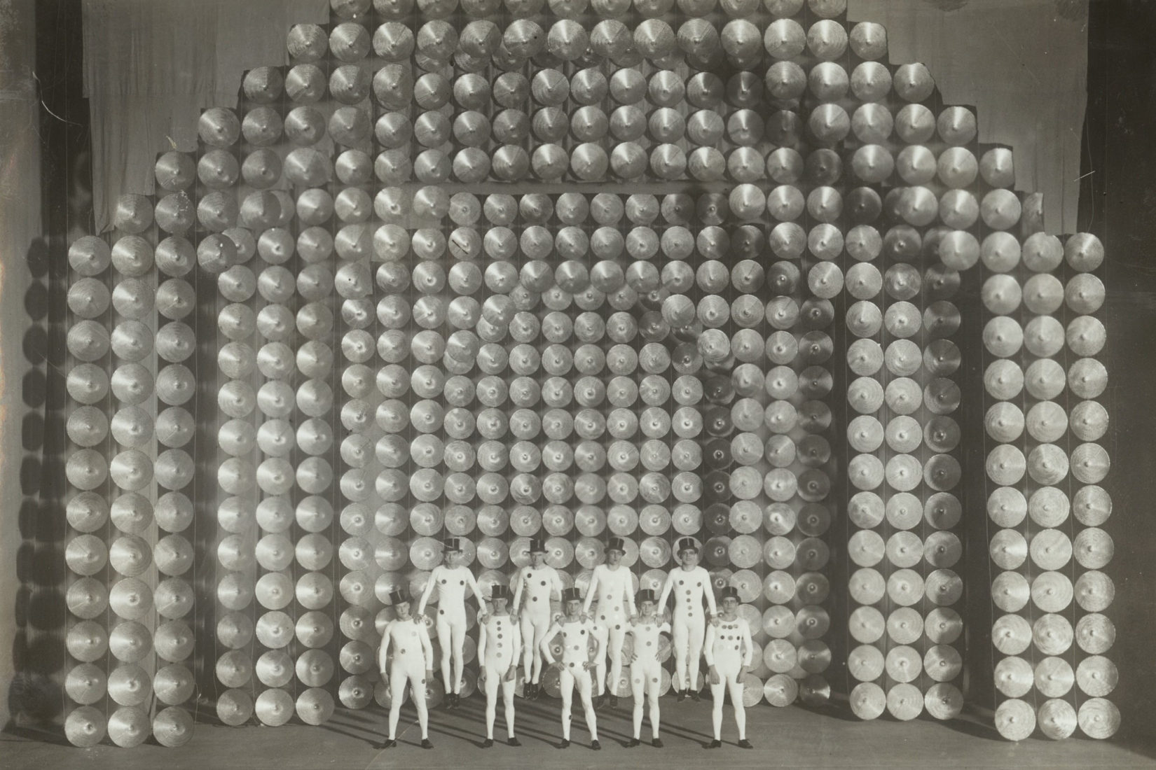 Black and white archive image of the Ballets suédois. Nine dancers stand in a formation in front of a stage set.