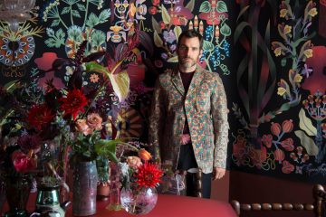 Pierre Marie se tient devant sa tapisserie Ras El Hanout, face à une table avec des vases remplis de fleurs.