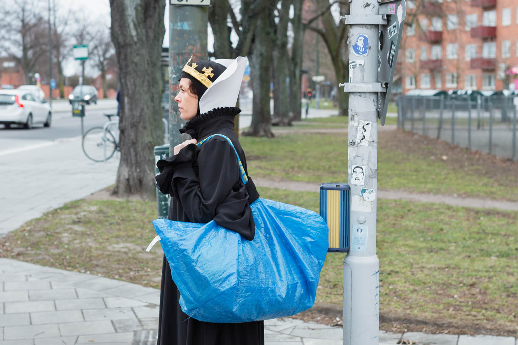 Liv Strömquist is standing in the street, showing her profile, in a long black dress, wearing a crown and a big blue Ikea bag on her shoulder.