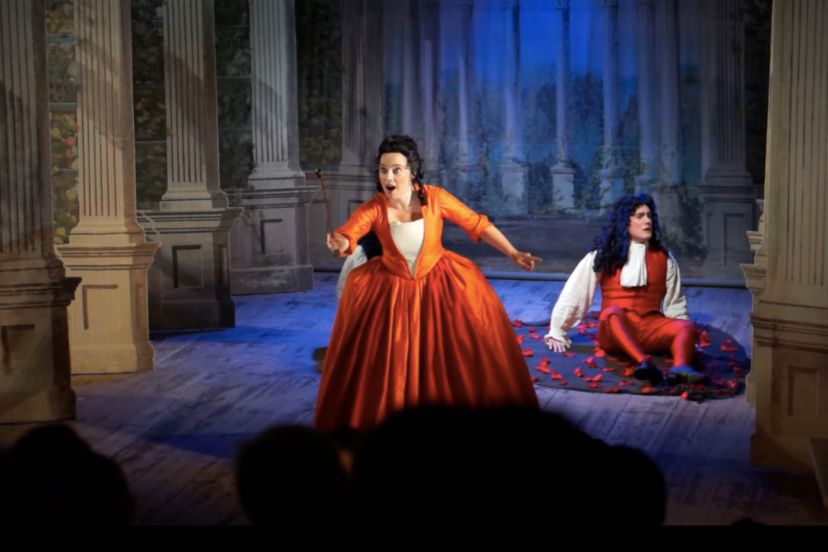 Annastina Malm, chanteuse d'opéra, chantant sur scène vêtue d'une robe d'époque orange, avec un homme assis sur le plancher en arrière-plan.
