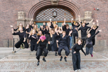 Les jeunes du Chœur de chambre du lycée Hvitfeldtska, tou.te.s vêtu.e.s de noir, sautent joyeusement dans les airs devant ce qui semble être le bâtiment du lycée.