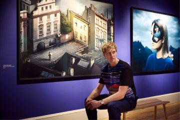 Photographer Erik Johansson is sitting on a bench in the exhibition room at Institut suédois, with his large scale photographs in the background.