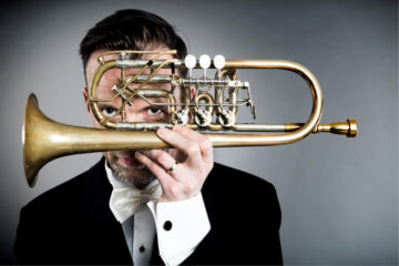 A musician holds a trumpet in front of his face and watches the camera through his instrument.
