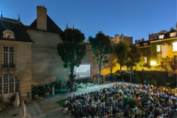 Photo of Institut suédois' garden at dusk, filled with people sitting on the grass in front of the giant screen on the brick wall.