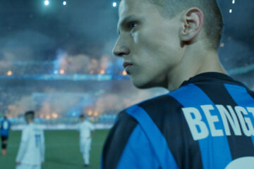 Photo du visage concentré d'un jeune joueur de football, de profil, avec en arrière-plan un match en cours dans un grand stade plein de monde.