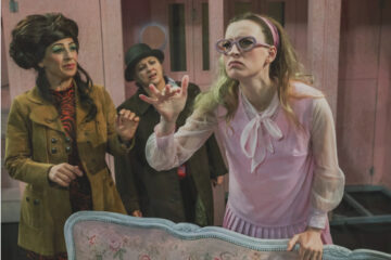 Trois comédiens sur scène dont l'une, au premier plan, vêtue d'une robe rose, semble concentrée sur un bruit, et les deux autres, derrière elle, semblent interrogatifs.