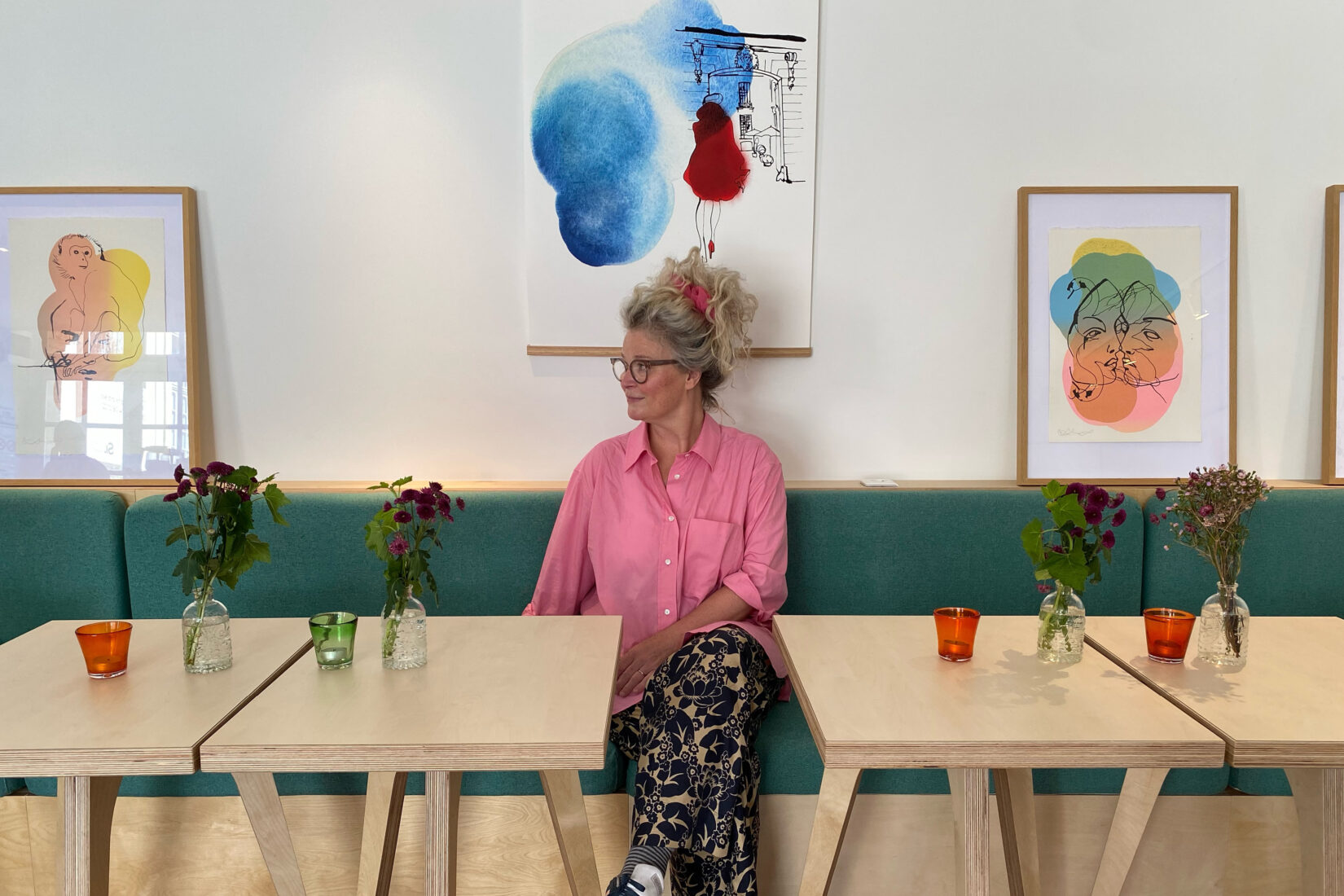 Stina Wirsén assise sur une banquette au café FIKA avec ses oeuvres derrière et à côté d'elle sur le mur.