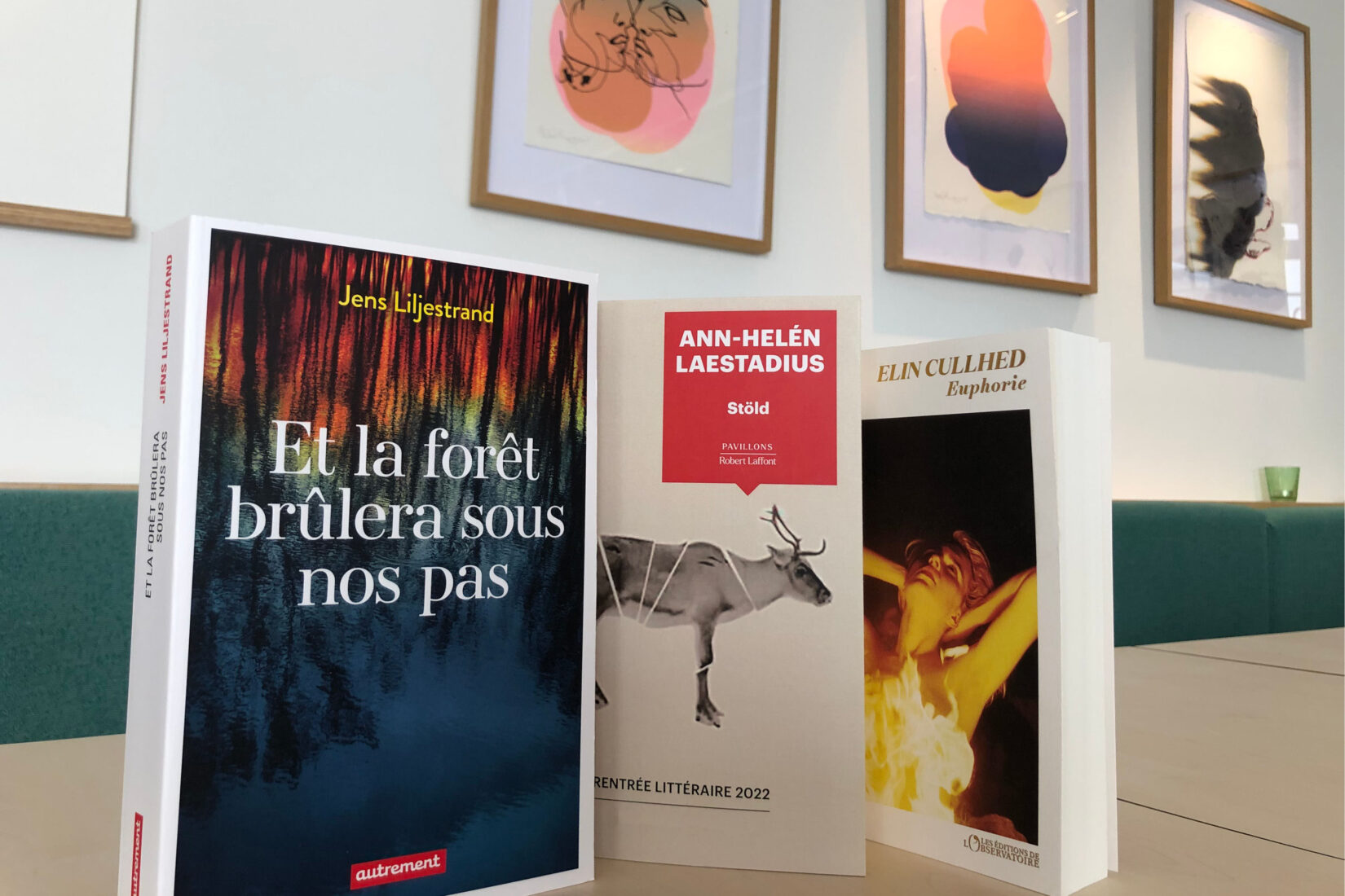 Photo des trois livres autour desquels se tient l'apéro littéraire, debout sur une table dans le café FIKA avec les œuvres de Stina Wirsén en arrière-plan.