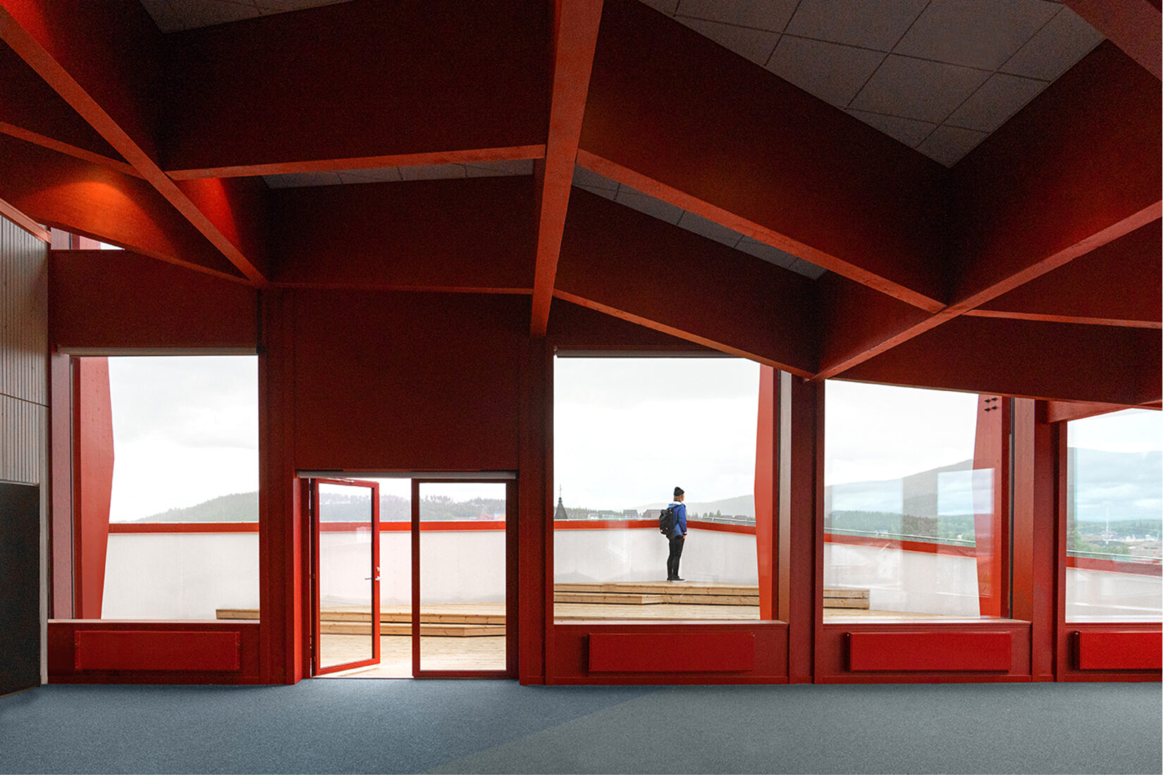 Photo prise de l'intérieur d'un bätiment moderne aux structures rouges, avec dans le fond une porte ouverte sur une terrasse et une personne qui se tient là pour admirer la vue.