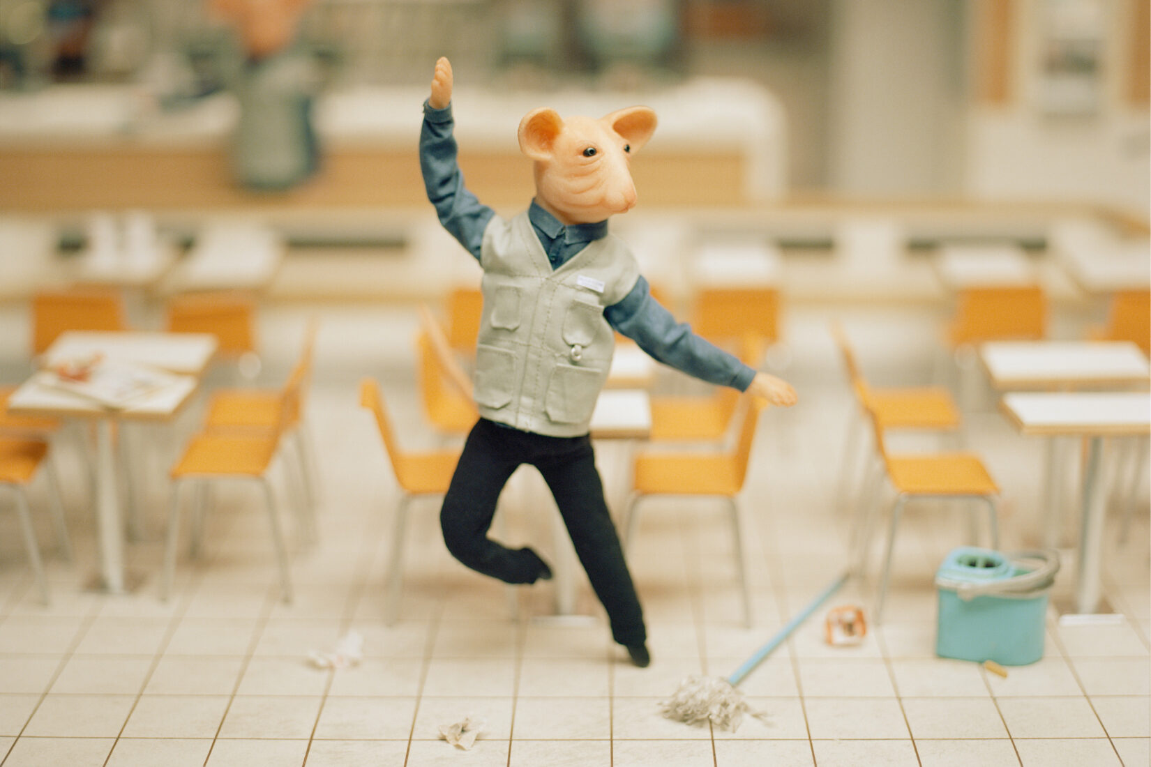 Photo de la maquette miniature d'une salle de cantine vide avec une figurine représentant une souris en train de danser avec à ses pieds un seau et un balai à serpillère abandonnés.