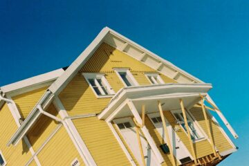 photo d'une maison en bois jaune qui bascule dans les airs, contre un ciel bleu.