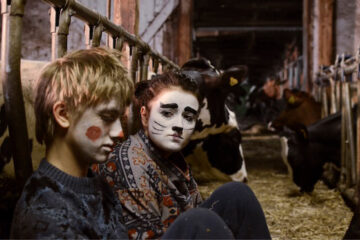 Photo of two characters sitting on the ground in a stable, one grimmed as a clown and the other one as a cat, both looking sad.