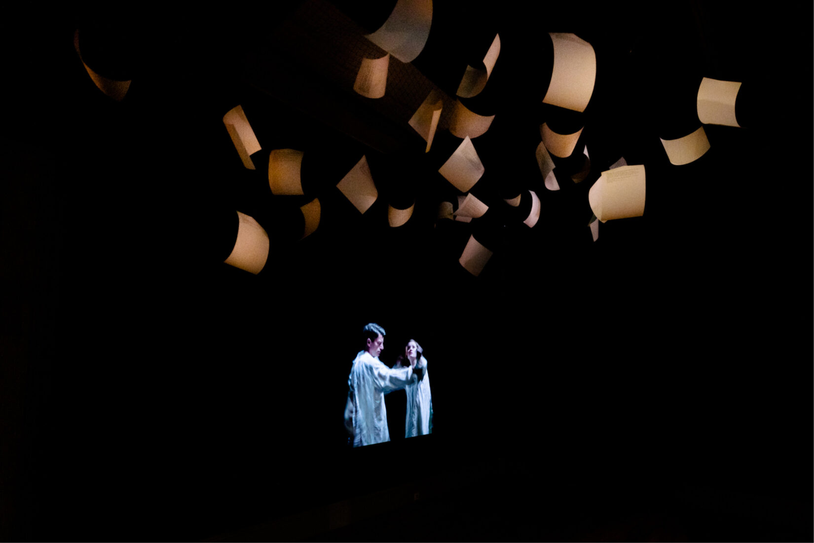Photo of manuscript pages flying from the ceiling in the dark, and image of two actors acting a scene in the background.