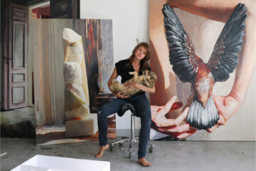 Photo of Sara-Vide Ericson with her big paintings in the background and holding a dog in her arms.