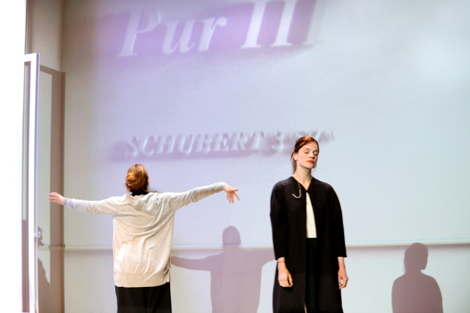 Photo d'une scène de la mise en scène de "Pur" à la Comédie-Française, montrant une femme debout de face les yeux fermés, et une femme de dos les bras étendus sur les côtés.