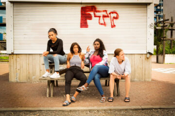 Four young women are sitting on a bench in front of a tagged white wall.