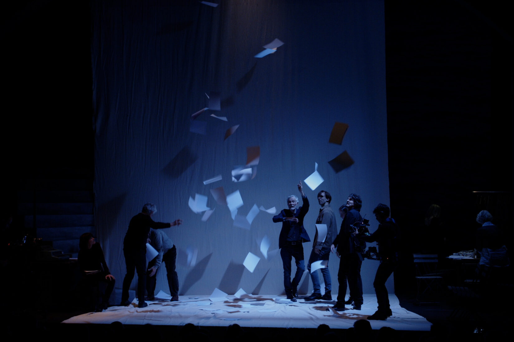 Dans une atmosphère bleu nuit, des comédiens sur scène tendent les bras vers des feuilles manuscrites qui tombent du ciel.
