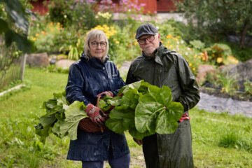 A couple of cultivators is standing in front of the camera, in their garden, holding vegetables.