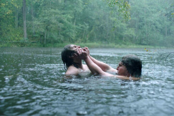 Deux personnes sont immergées dans un lac ou une rivière, sous la pluie, en train de se battre.