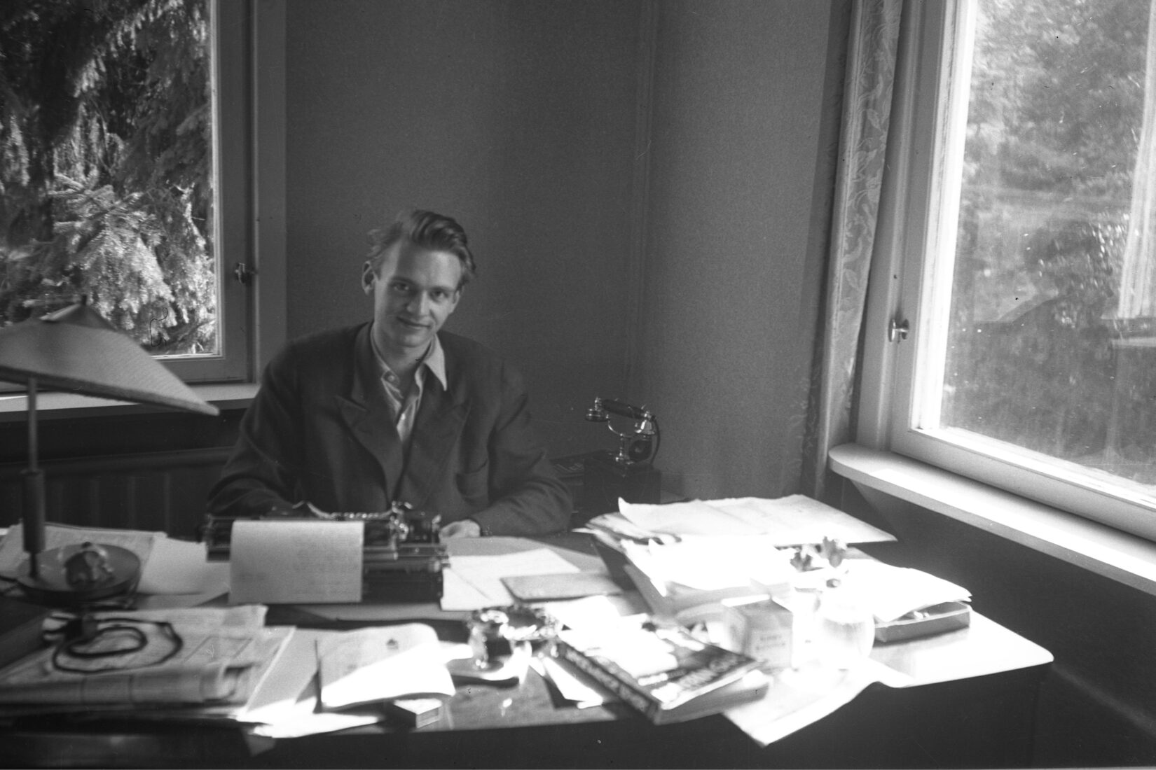 Photo en noir et blanc de Stig Dagerman assis à son bureau, entouré de fenêtres par lesquelles on voit des arbres, souriant.