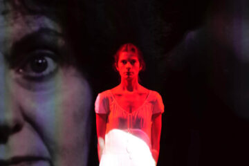une jeune femme se tient debout et regarde droit devant elle. la projection d'un visage plus agée est projeté derrière elle et une lumière rouge la recouvre