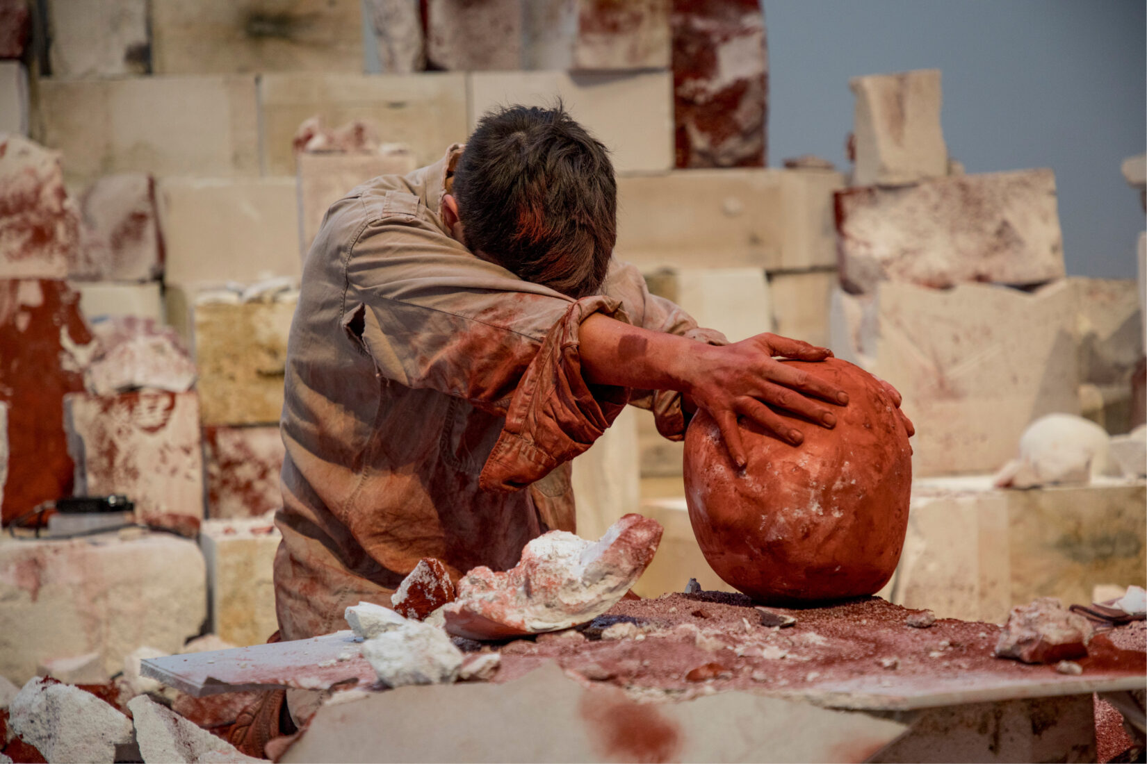 Photo de l'artiste concentrée sur sa performance, tenant une boule couverte d'encre rouge à bout de bras, l'installation de béton en arrière-plan.
