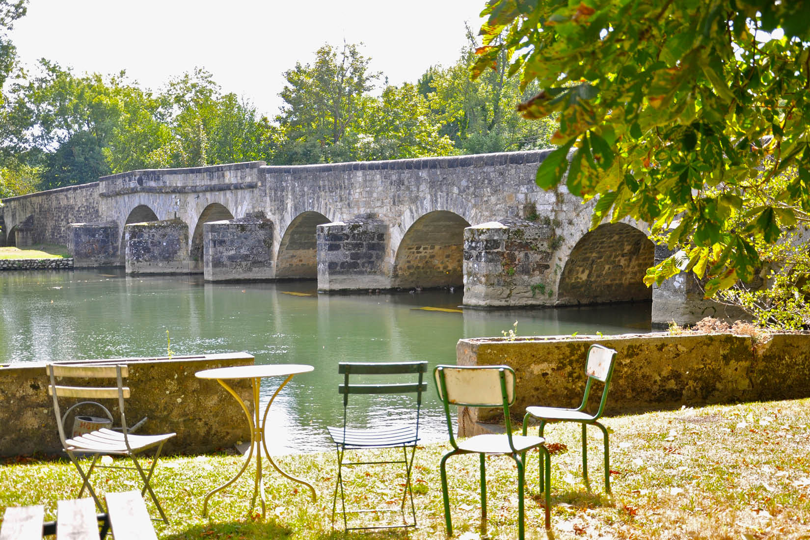 Quelques chaises et une table ronde disposées au bord d'une rivière, près d'un pont en pierre.