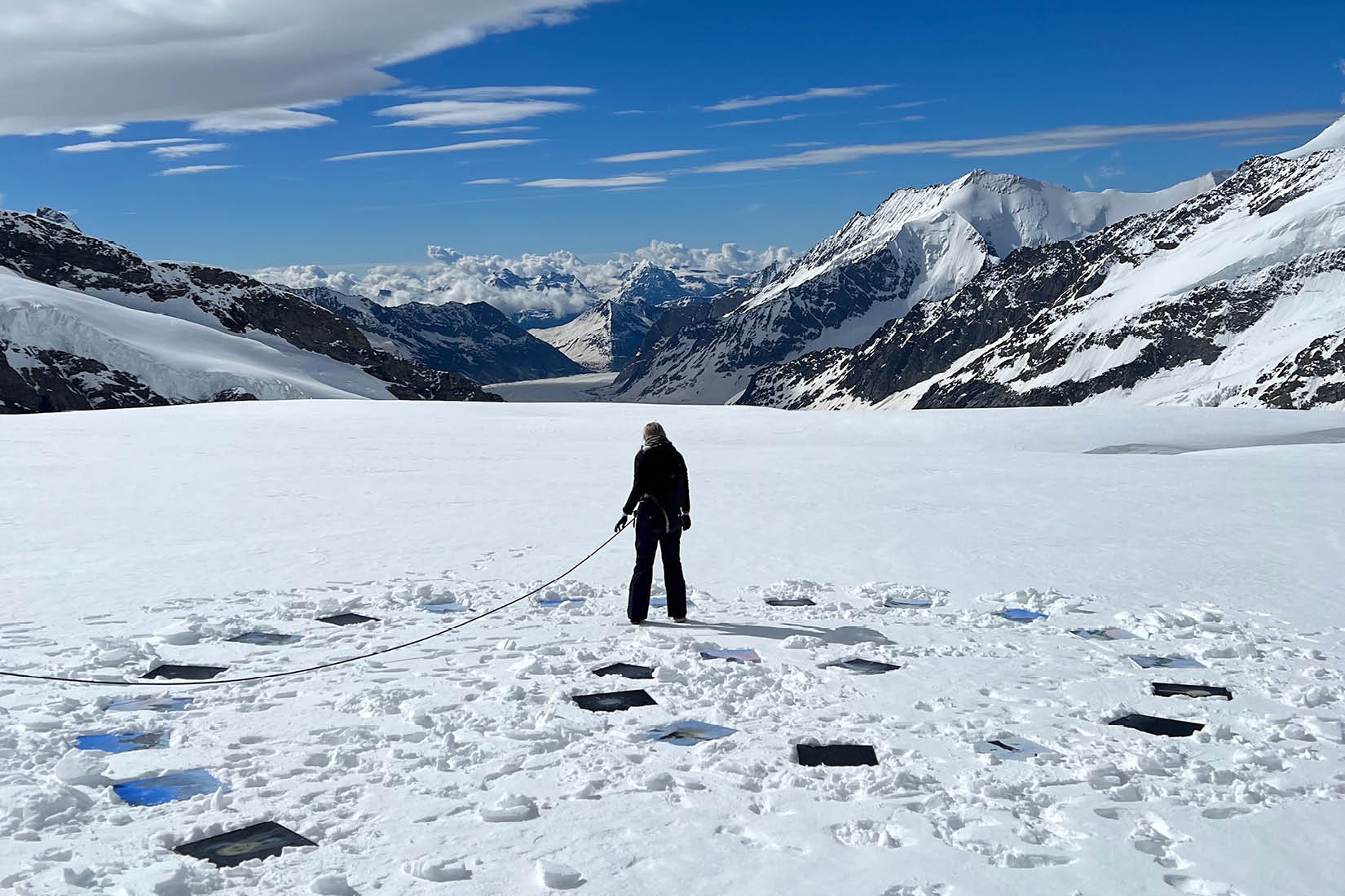 Une personne se tient debout dans la neige, dans un paysage de montagnes, des oeuvres d'art disposées en spirale autour d'elle.