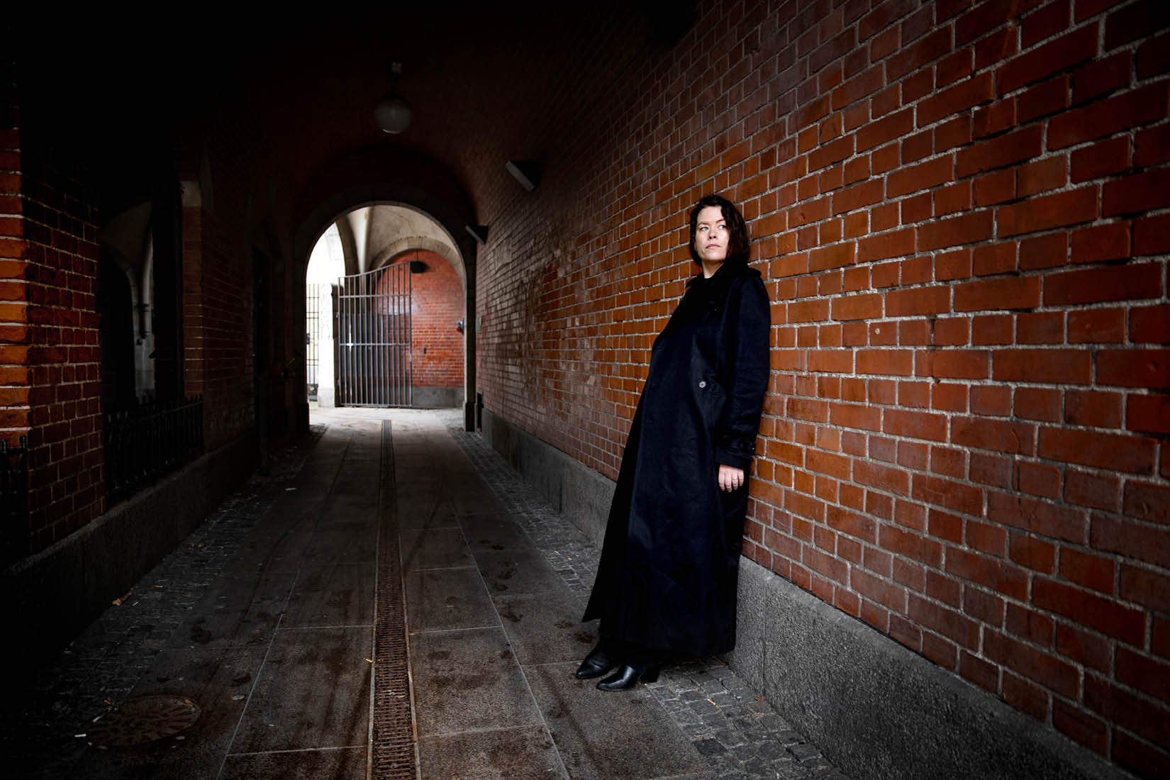 Photo de Liv Strömqvist, longue robe noire et visage encadré de cheveux noirs, se tenant contre un mur en brique dans ce qui ressemble à un tunnel.