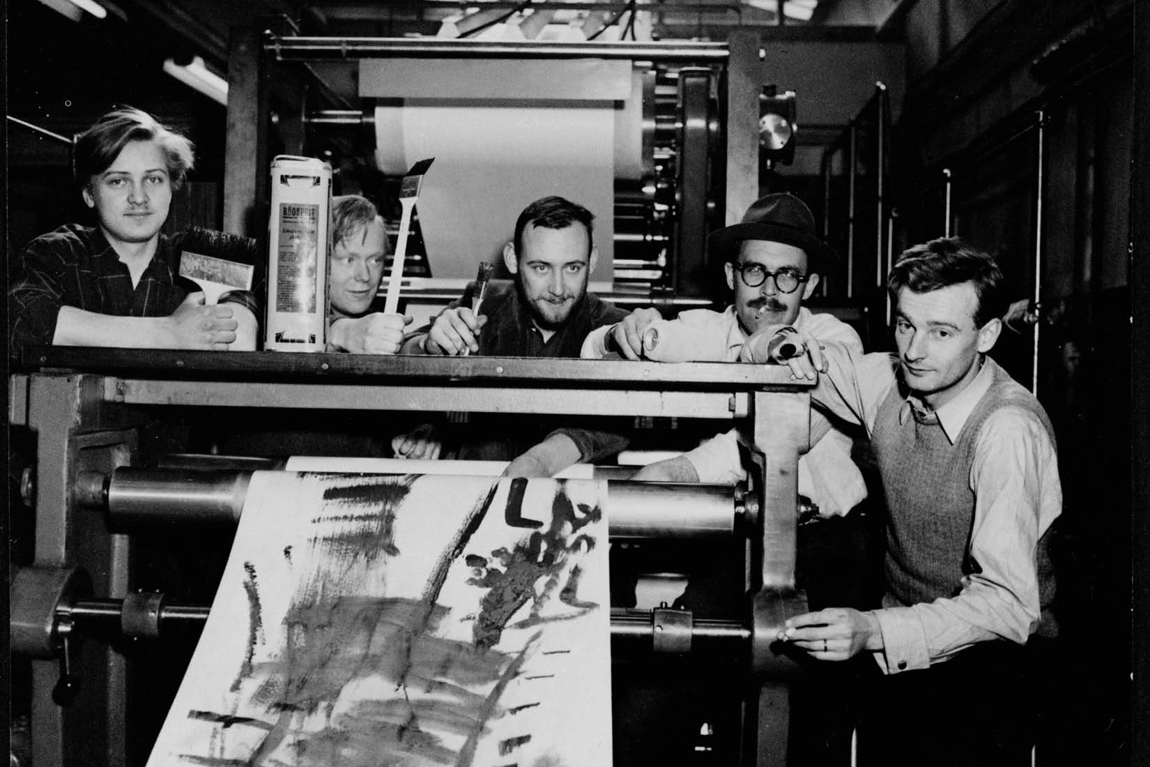 Photo en noir et blanc montrant quatre hommes derrière une presse, avec une grande affiche en cours d'impression.