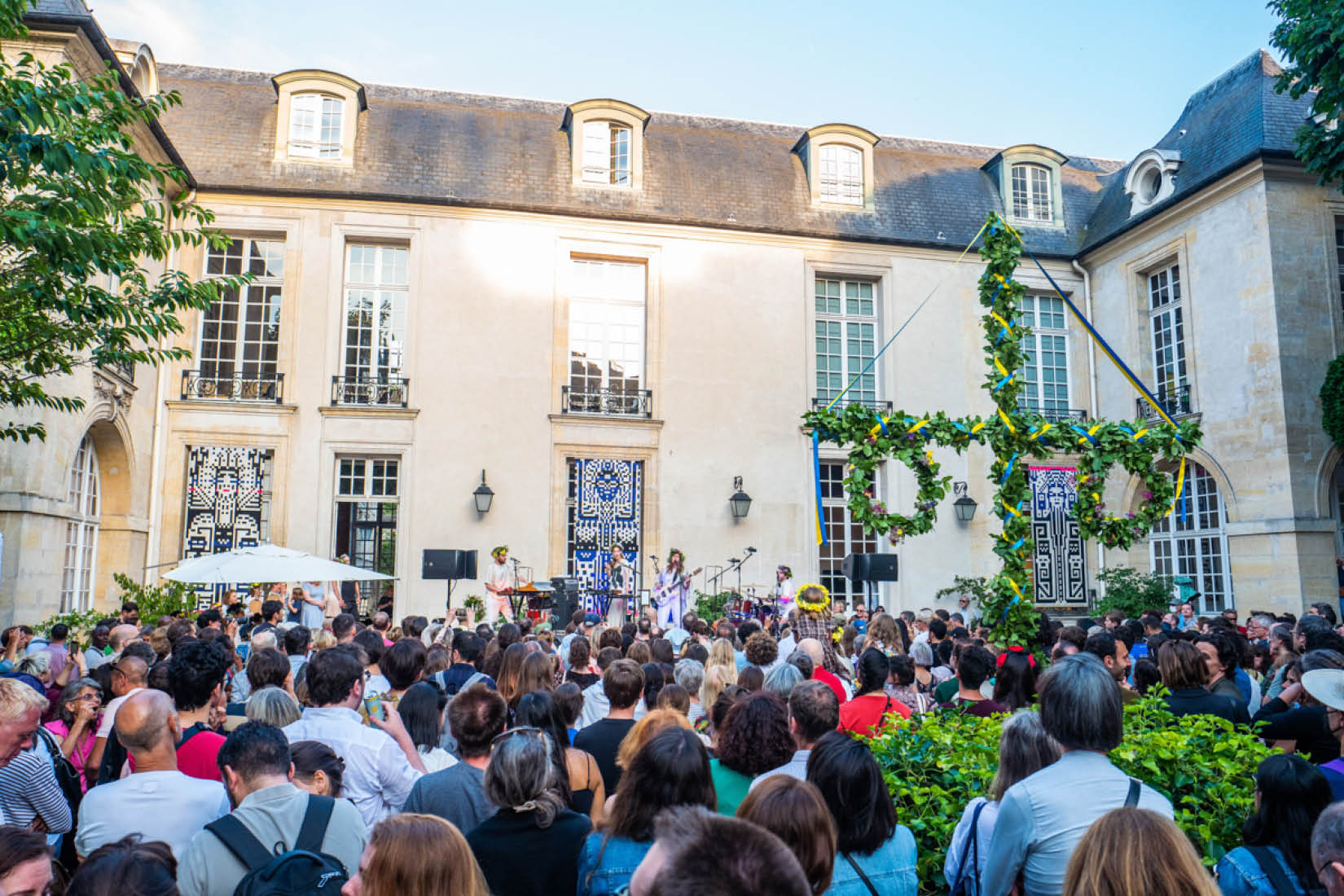 Foule rassemblée devant une scène et autour d'un mât fleuri dans le jardin de l'Institut suédois.