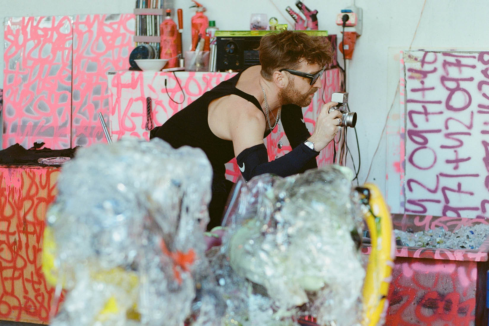 Artiste tenant un appareil photo au milieu d'œuvres en verre entourées d'inscriptions roses.