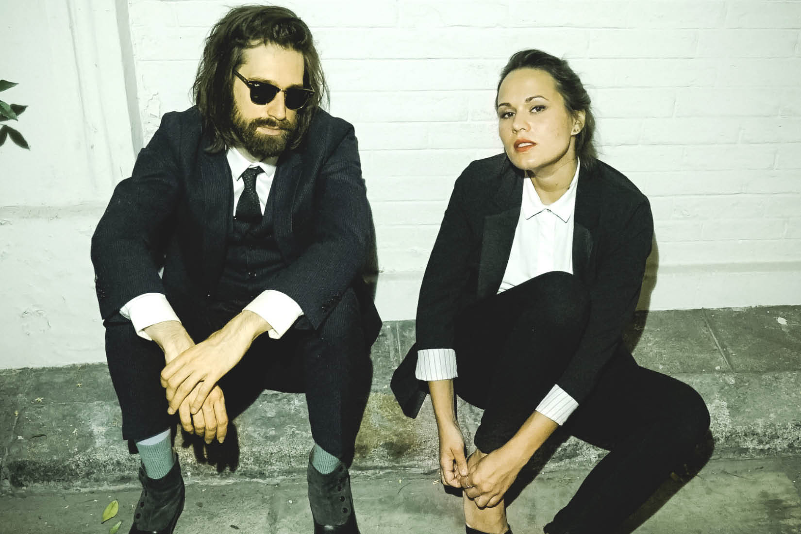 Un homme et une femme en costume noir et chemise blanche assis sur un trottoir, côte-à-côte.