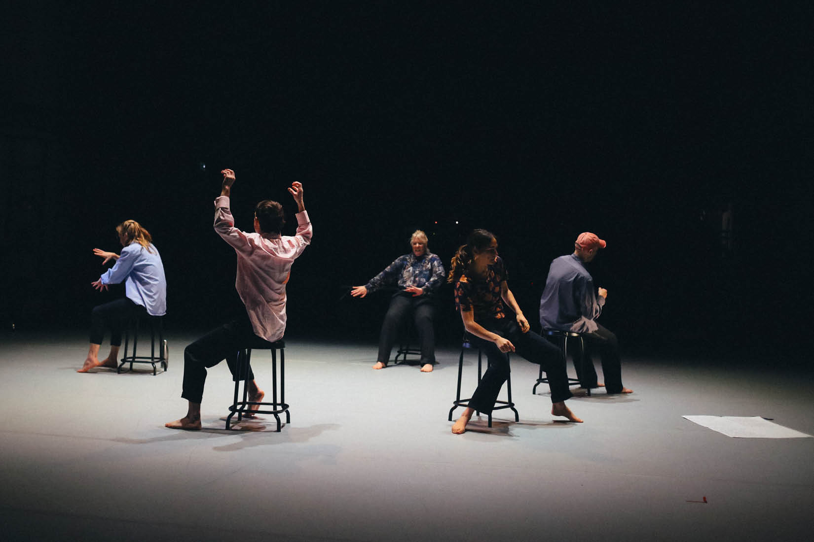 Cinq danseurs sur scène, assis en cercle sur des tabourets, faisant des mouvements de bras.