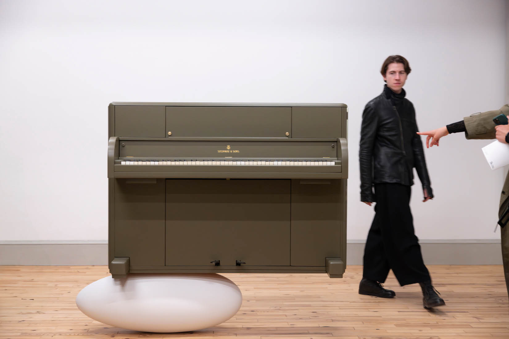 A piano on a white cocoon in a white room, with people around.